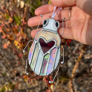 May include: A stained glass ornament shaped like a beetle with a red heart in the center. The beetle is iridescent with white, pink, and purple colors.