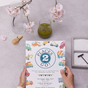 May include: A colorful invitation card with a blue circle and the text "Places 2 Go!" surrounded by illustrations of vehicles. The text "Please join us in celebrating as Jackson turns two" is below the circle. The date and time of the event are listed as "December 16th, 2023, 4pm - 6pm". The address is "4444 N Street Ave, City, ST 12345". The RSVP information is "Please RSVP to Rachelle at 333-777-7777".
