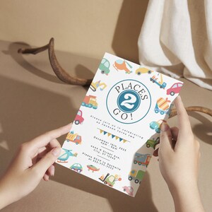 May include: A white invitation card with a blue circle that says "Places 2 Go!" and has a number 2 in the center. The card is decorated with colorful illustrations of vehicles, including trucks, cars, helicopters, and construction equipment. The text on the card reads "Please join us in celebrating as Jackson turns two. December 16th, 2023, 4pm - 6pm, 4444 N Street Ave., City, ST 12345. Please RSVP to Rachelle at 333-777-7777."