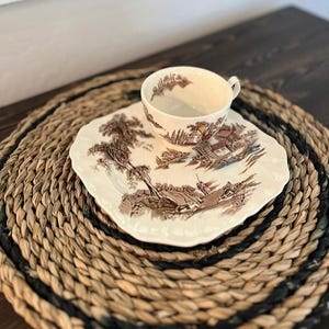 May include: A vintage teacup and saucer set with a brown landscape design. The teacup sits on a square saucer, both with a cream background and brown transferware images. The set rests on a round, woven placemat with black accents.