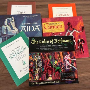 May include: A collection of vintage opera librettos, including "Aida" by Giuseppe Verdi and "Carmen" by Georges Bizet. The covers feature colorful illustrations and text, with titles like "The Tales of Hoffmann" and "The Metropolitan Opera Record Club."