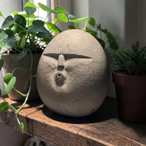 May include: A smooth, egg-shaped stone sculpture featuring a carved face with narrow, horizontal eyes and a small, circular, pursed mouth. The sculpture has a matte, light grey, textured surface and sits upright as a standalone decorative object.