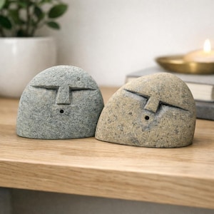 Puede incluir: Dos porta-inciensos de piedra en forma de cúpula con rasgos faciales tallados. Uno es gris y el otro es beige. Cada piedra tiene una línea horizontal para los ojos y una vertical para la nariz, con un pequeño orificio circular debajo.