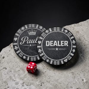 May include: Two circular black poker chips with white engraved details. One chip features the word DEALER and a star, while the other displays the name Paul, a crown, and a star. Both chips have card suit symbols along the edge. A single red die is included.
