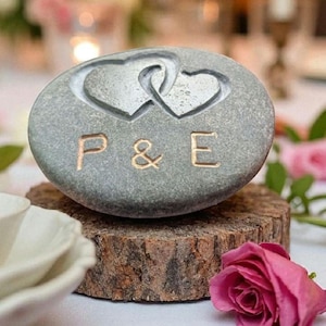 May include: A smooth, oval-shaped grey river stone featuring two interlocking heart shapes carved into the top surface. Below the hearts, the letters P, &, and E are engraved in a clean, sans-serif font, creating a personalised decorative object.