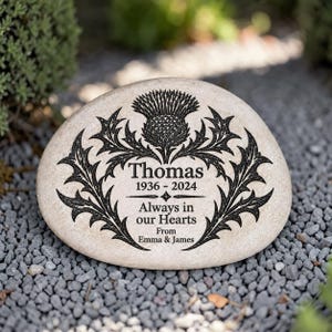 May include: A light-colored, smooth, oval memorial stone featuring a black engraved thistle design. The text reads: Thomas, 1936 - 2024, Always in our Hearts, From Emma & James. The engraving is centered on the stone surface.