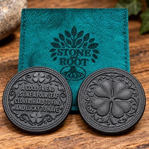 Peut inclure: Deux jetons noirs circulaires avec des motifs en relief placés devant une pochette en velours bleu sarcelle. Un jeton présente un trèfle à quatre feuilles, tandis que l'autre affiche le texte : A GOOD FRIEND IS LIKE A FOUR LEAF CLOVER.