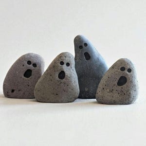 Peut inclure: Quatre pierres grises et lisses disposées en groupe, chacune présentant deux petits yeux circulaires et une bouche ovale peints en noir. Les pierres varient en taille et en forme, avec une pierre plus haute et plus sombre placée derrière.