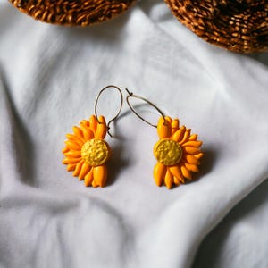 Könnte beinhalten: Ein Paar goldene Creolen mit gelben Sonnenblumen-Anhängern. Die Ohrringe liegen auf einem weißen Stoffhintergrund.