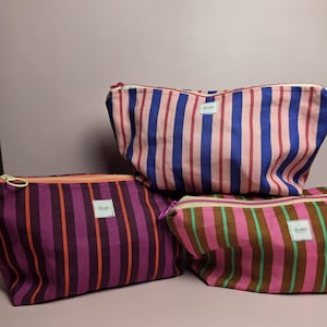Op de afbeelding: Drie etuis met rits en verticale strepen in verschillende kleurcombinaties. Eén etui heeft roze en blauwe strepen, een andere paarse en oranje strepen, en de derde heeft roze, bruine en groene strepen. Elk etui heeft een klein wit label.