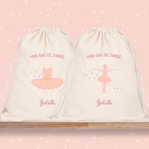 Könnte beinhalten: Zwei Canvas-Kordelzugbeutel mit Ballettdesign. Eine Tasche zeigt einen rosa Tütü und den Text "MON SAC DE DANSE" und "Juliette". Die andere Tasche hat eine Ballerina-Grafik und denselben Text.