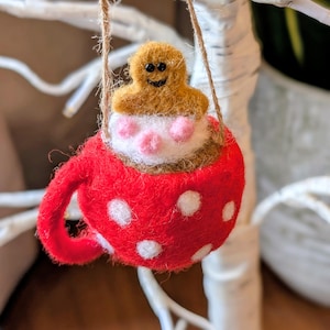 May include: A festive felt Christmas ornament. It depicts a gingerbread man in a red mug with white polka dots, filled with white and pink whipped cream. The ornament hangs from a twine string, ideal for festive decorations.