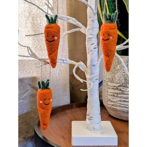 May include: Three orange felt carrot decorations with green tops and smiling faces hang from a decorative white tree. The carrots are suspended by twine and have pink cheeks. The tree is on a wooden table.