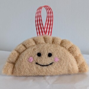 May include: A felt dumpling decoration with a smiling face, black eyes, and pink cheeks. The beige dumpling has a red and white gingham ribbon for hanging. A cute, handmade food-themed ornament.