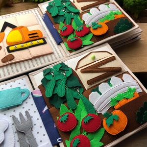 May include: A handmade felt quiet book with various garden-themed pages. The pages feature colorful felt vegetables like tomatoes, cucumbers, and carrots, along with a felt excavator and other interactive elements. The book is designed for children.