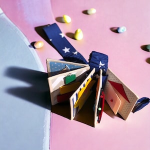 May include: A blue and white fabric baby book with a variety of textures and colors. The book is open to reveal different pages with a yellow sun, a red triangle, and a green square.
