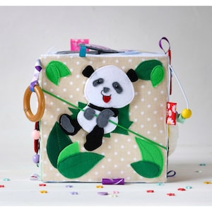 Può includere: Un cubo di stoffa beige con un'applicazione di orso panda. Il panda è bianco con dettagli neri e braccia e gambe grigie. Foglie di feltro verdi e un anello di legno sono attaccati. Il cubo ha vari attacchi colorati.