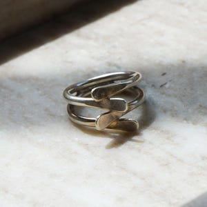 May include: A silver-colored ring with a unique, layered design. The ring features a spiral shape with three bands, each with a stylized, curved element. The ring is set against a light-colored, textured surface.