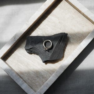 May include: A silver ring with a unique, minimalist design rests on a dark gray, textured stone. The stone is displayed in a rectangular, light-colored tray, set against a neutral linen background. The ring's design features a double loop with a cross detail.