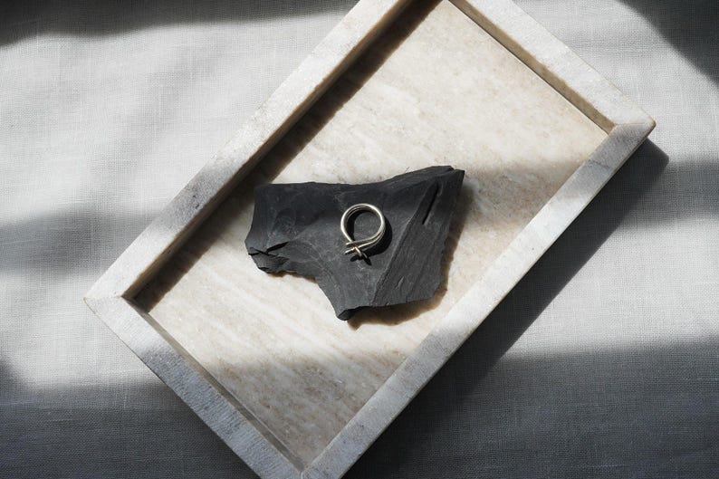 May include: A silver ring with a unique, minimalist design rests on a dark gray, textured stone. The stone is displayed in a rectangular, light-colored tray, set against a neutral linen background. The ring's design features a double loop with a cross detail.