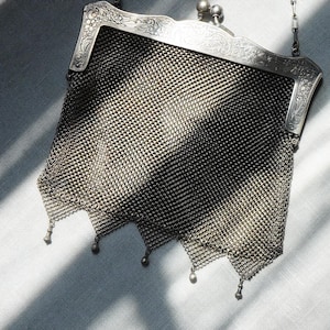 May include: A vintage silver mesh handbag with a decorative frame and chain handle. The bag features a fine, woven mesh design with a scalloped bottom edge. The frame is detailed with ornate floral patterns.
