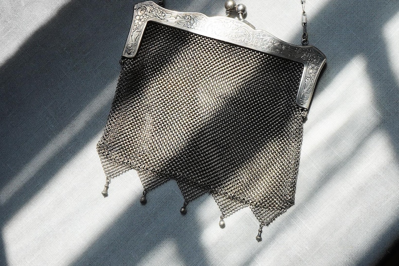 May include: A vintage silver mesh handbag with a decorative frame and chain handle. The bag features a fine, woven mesh design with a scalloped bottom edge. The frame is detailed with ornate floral patterns.