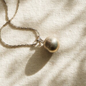 Puede incluir: Un collar plateado con un colgante redondo y dorado. La cadena tiene un dise&ntilde;o delicado y texturizado. La joya se encuentra sobre un fondo neutro y texturizado, con sombras que a&ntilde;aden profundidad.