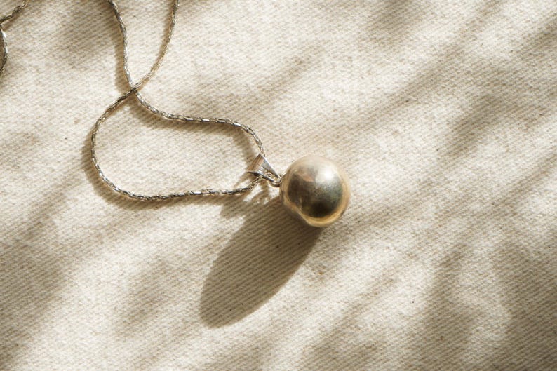 Puede incluir: Un collar plateado con un colgante redondo y dorado. La cadena tiene un dise&ntilde;o delicado y texturizado. La joya se encuentra sobre un fondo neutro y texturizado, con sombras que a&ntilde;aden profundidad.