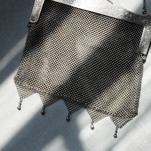 May include: An antique silver mesh handbag with a decorative frame. The bag features a delicate chainmail design and small dangling embellishments at the bottom. The frame has ornate detailing.
