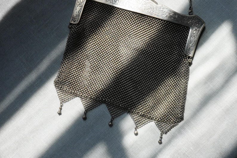 May include: An antique silver mesh handbag with a decorative frame. The bag features a delicate chainmail design and small dangling embellishments at the bottom. The frame has ornate detailing.