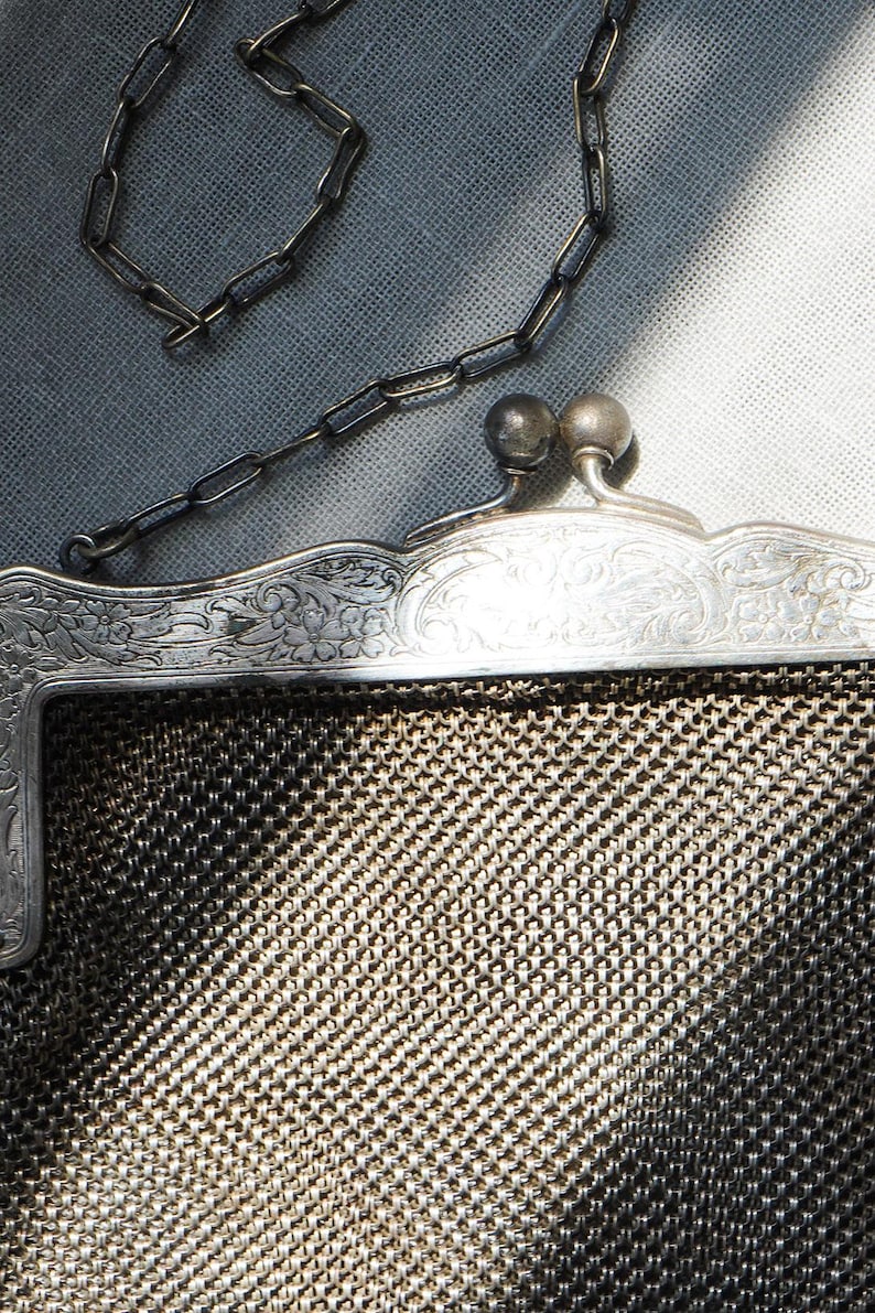 May include: Close-up of a vintage silver mesh purse with a chain handle. The purse has a decorative floral design on the frame and a clasp with two round ball details. The mesh is a fine, woven pattern.