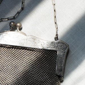 May include: An antique silver mesh purse with a decorative frame and chain handle. The frame features ornate floral and scrollwork designs. The purse's mesh body is a dark, metallic color, and the chain is silver.