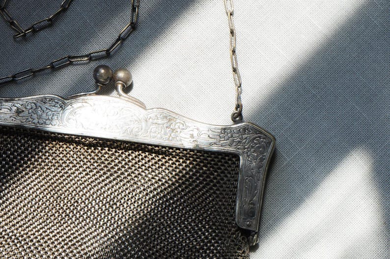 May include: An antique silver mesh purse with a decorative frame and chain handle. The frame features ornate floral and scrollwork designs. The purse's mesh body is a dark, metallic color, and the chain is silver.