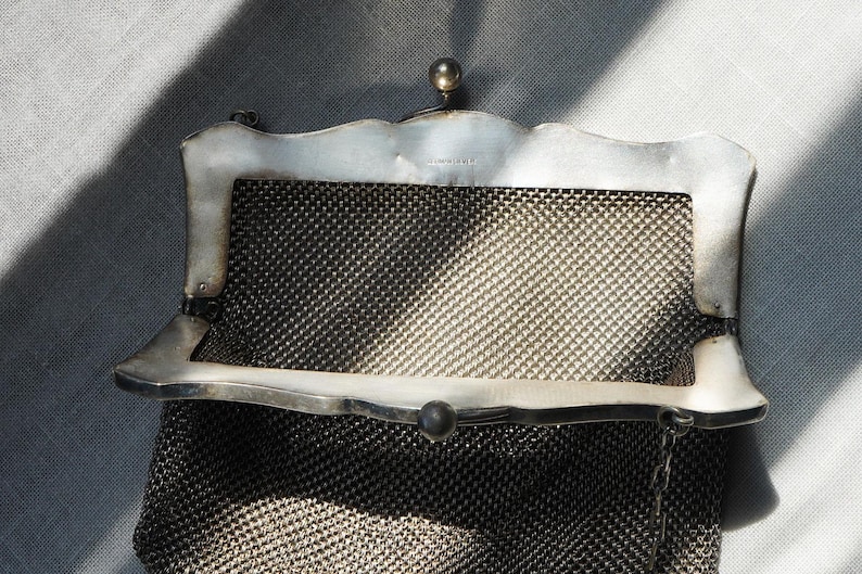 May include: A vintage silver mesh purse with a decorative frame. The purse features a fine, woven mesh body and a silver frame with a scalloped edge. A small, round clasp secures the purse.