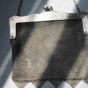 May include: An antique silver mesh purse with a decorative frame and chain handle. The purse features a detailed woven mesh body, ornate floral detailing on the frame, and small dangling charms at the bottom. The purse is a vintage accessory.