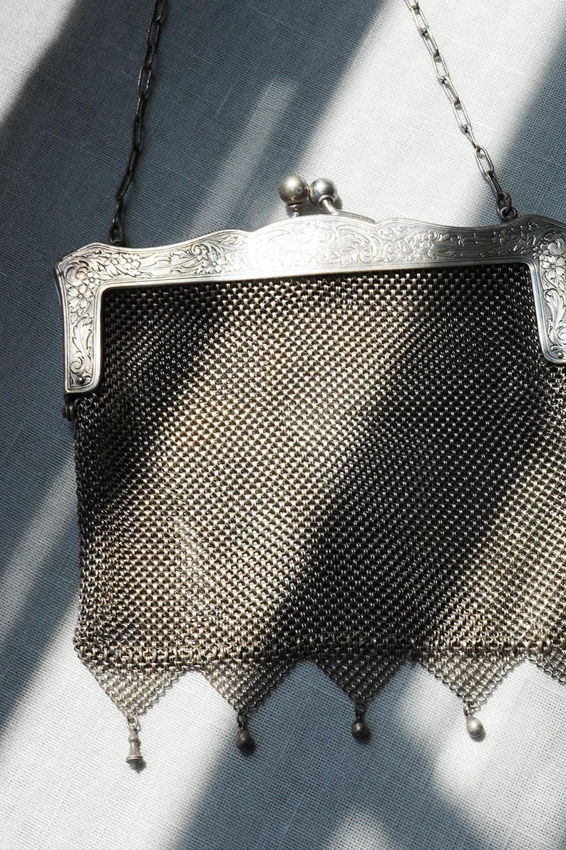 May include: An antique silver mesh purse with a decorative frame and chain handle. The purse features a detailed woven mesh body, ornate floral detailing on the frame, and small dangling charms at the bottom. The purse is a vintage accessory.