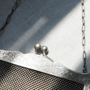 May include: An antique silver mesh purse with a decorative floral frame. The purse has a chain handle and a clasp with two small silver spheres. The mesh is a dark gray color.