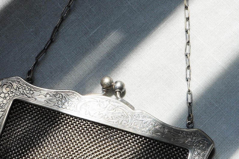 May include: An antique silver mesh purse with a decorative floral frame. The purse has a chain handle and a clasp with two small silver spheres. The mesh is a dark gray color.
