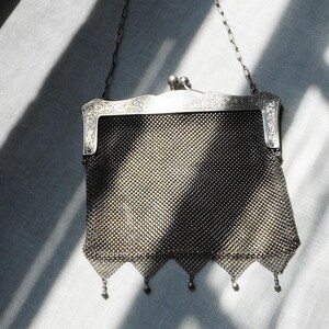May include: An antique silver mesh purse with a decorative frame and chain handle. The purse features a fine, woven mesh body and small dangling beads along the bottom edge. The frame has ornate floral detailing.