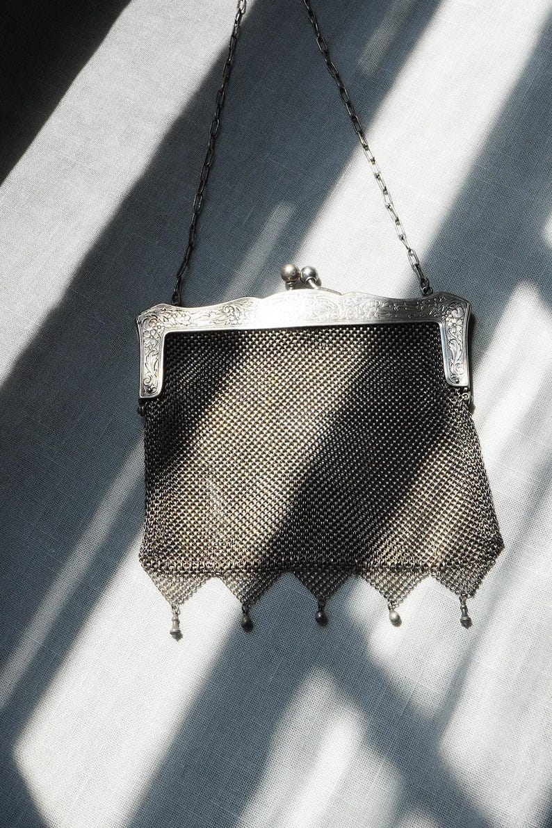 May include: An antique silver mesh purse with a decorative frame and chain handle. The purse features a fine, woven mesh body and small dangling beads along the bottom edge. The frame has ornate floral detailing.