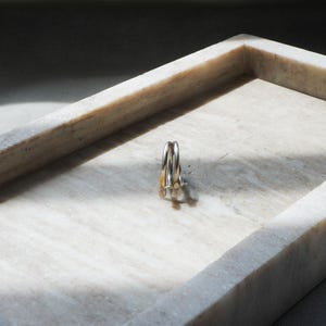 May include: A set of three rings, two silver and one gold, resting inside a rectangular marble tray. The tray is white with gray veining, and the rings are positioned in the center, casting a shadow.