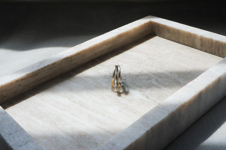 May include: A set of three rings, two silver and one gold, resting inside a rectangular marble tray. The tray is white with gray veining, and the rings are positioned in the center, casting a shadow.