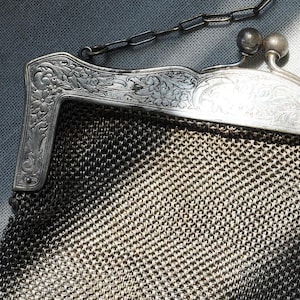 May include: Close-up of a vintage silver mesh purse. The purse features an ornate, engraved silver frame with floral detailing and a delicate chain handle. The mesh body has a woven pattern.