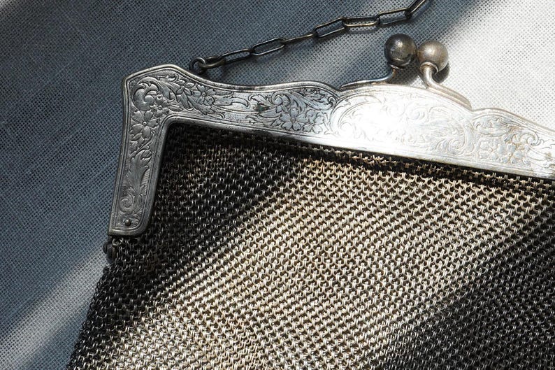 May include: Close-up of a vintage silver mesh purse. The purse features an ornate, engraved silver frame with floral detailing and a delicate chain handle. The mesh body has a woven pattern.