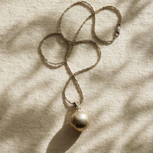 Puede incluir: Un collar de plata con un colgante redondo. La cadena es delicada y el colgante tiene una forma esf&eacute;rica lisa. El collar se muestra sobre una superficie texturizada de color blanquecino, con sombras.