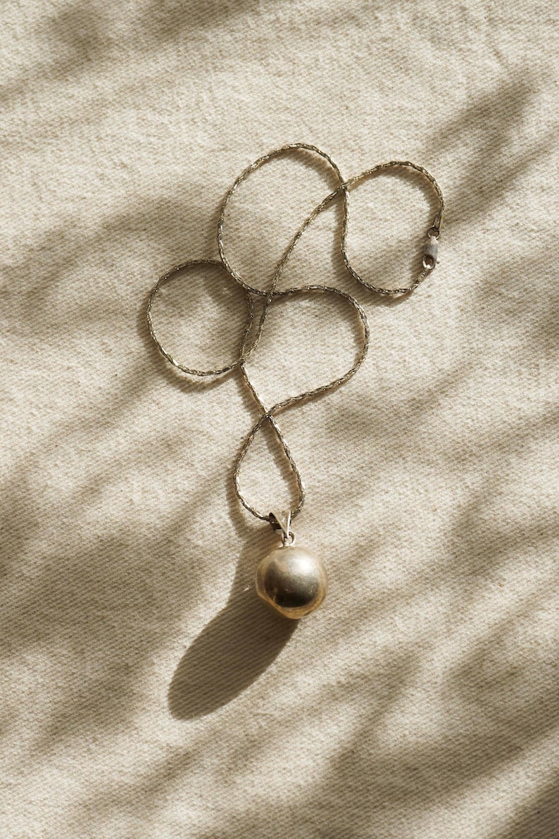 Puede incluir: Un collar de plata con un colgante redondo. La cadena es delicada y el colgante tiene una forma esf&eacute;rica lisa. El collar se muestra sobre una superficie texturizada de color blanquecino, con sombras.