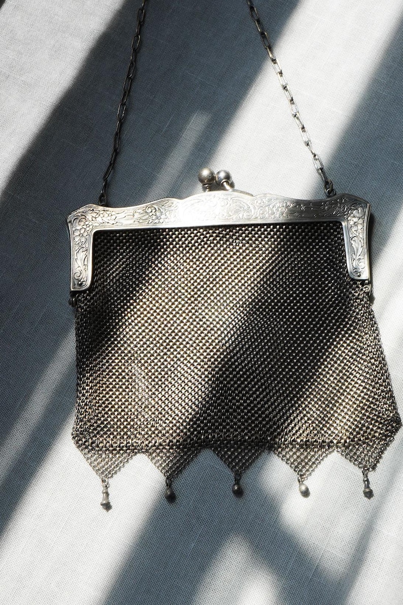 May include: A vintage silver mesh purse with a decorative frame and chain handle. The purse features a detailed floral design on the frame and small dangling ornaments at the bottom. The mesh is a fine, woven pattern.