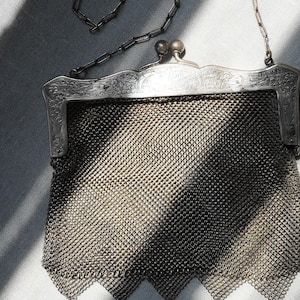 May include: An antique silver mesh purse with a decorative frame and chain handle. The purse features a detailed, ornate design on the frame and a delicate chain strap. The mesh body of the purse has a diamond pattern and is finished with small dangling charms.