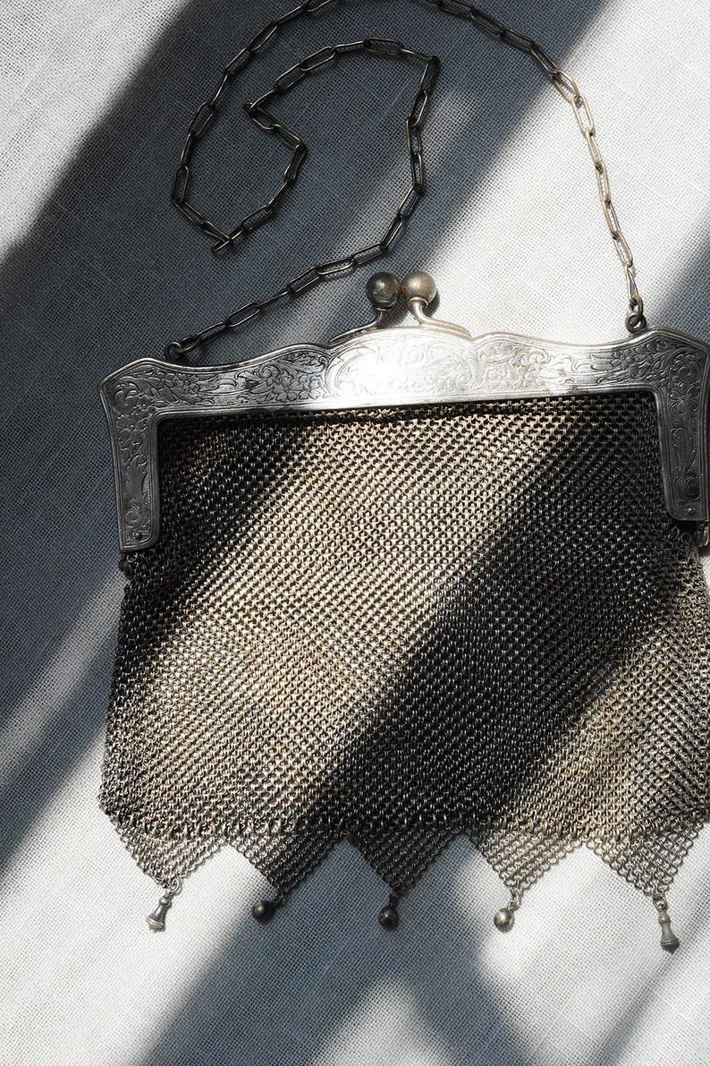 May include: An antique silver mesh purse with a decorative frame and chain handle. The purse features a detailed, ornate design on the frame and a delicate chain strap. The mesh body of the purse has a diamond pattern and is finished with small dangling charms.