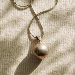Puede incluir: Un collar plateado con un colgante redondo. El colgante tiene una forma esf&eacute;rica lisa. La cadena tiene un dise&ntilde;o retorcido. El collar se muestra sobre una superficie clara y texturizada.